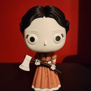 May include: A stylized vinyl figure with a large white head, black hair, and large round eyes. The figure wears a long, muted orange dress with white trim and holds a small white and black axe in its hands. The design features a smooth, matte finish.