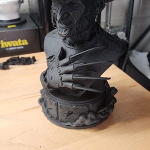 May include: A matte black 3D printed bust of a character wearing a fedora hat, featuring a scarred face and a glove with four long, sharp blades extending from the fingers. The figure is mounted on a circular base with a wood-plank texture.