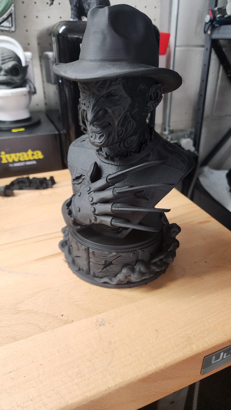 May include: A matte black 3D printed bust of a character wearing a fedora hat, featuring a scarred face and a glove with four long, sharp blades extending from the fingers. The figure is mounted on a circular base with a wood-plank texture.