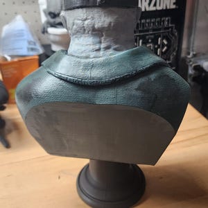 May include: A gray, textured bust of a figure wearing a dark green jacket and a black strap across the head. The sculpture features a detailed, weathered skin texture on the back of the head and neck, mounted on a solid, circular black pedestal base.