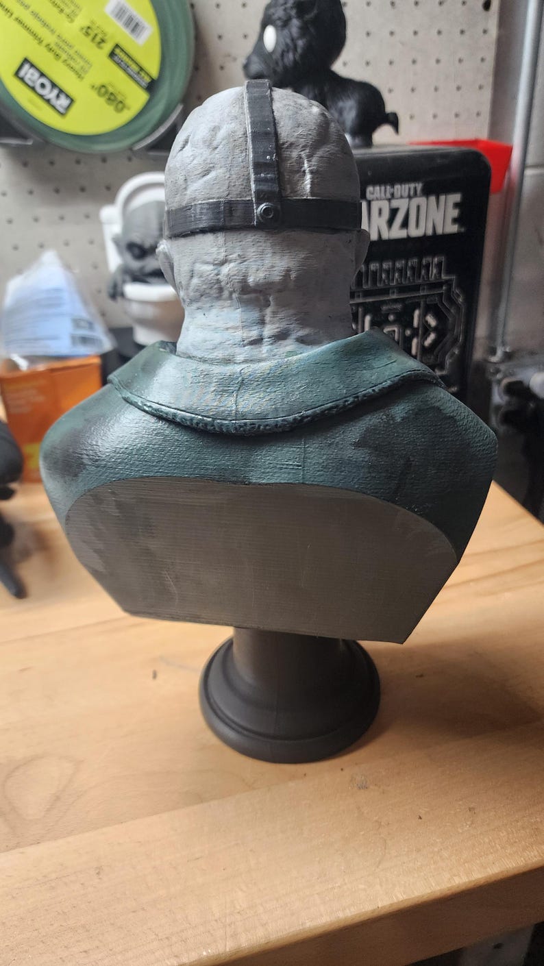 May include: A gray, textured bust of a figure wearing a dark green jacket and a black strap across the head. The sculpture features a detailed, weathered skin texture on the back of the head and neck, mounted on a solid, circular black pedestal base.