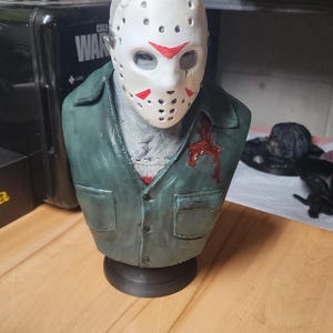 May include: A sculpted bust of a figure wearing a white hockey mask with red triangular markings and a weathered green collared shirt. The shirt features a dark red blood-like stain on the left chest area. The figure is mounted on a black base.