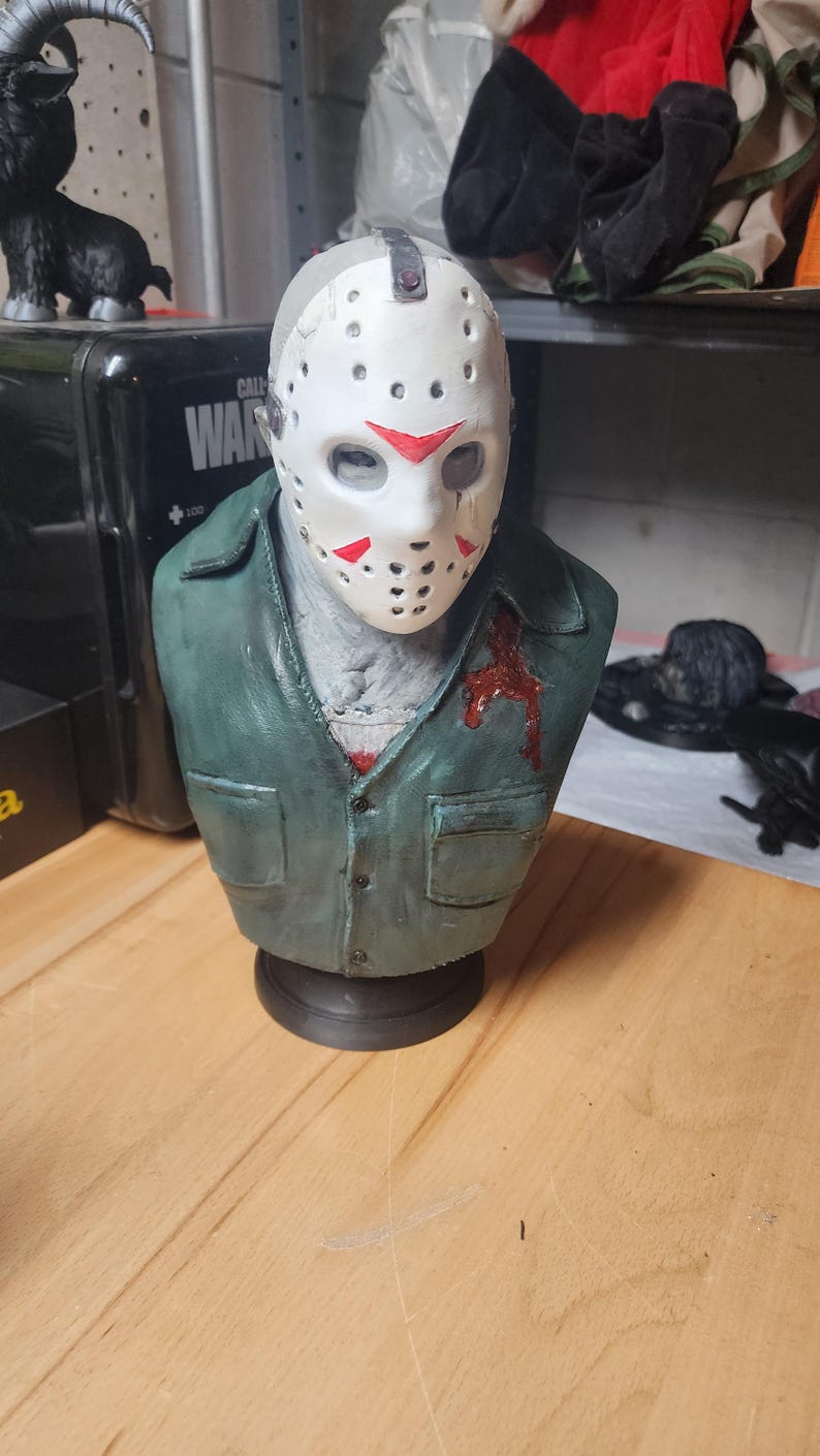 May include: A sculpted bust of a figure wearing a white hockey mask with red triangular markings and a weathered green collared shirt. The shirt features a dark red blood-like stain on the left chest area. The figure is mounted on a black base.