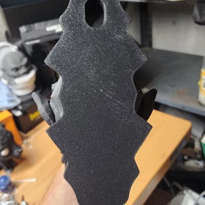 May include: A black, textured, vertically oriented object with a jagged, serrated edge profile and a single oval-shaped hole near the top. The surface has a matte, granular finish, and the overall shape is elongated with a pointed top and bottom.