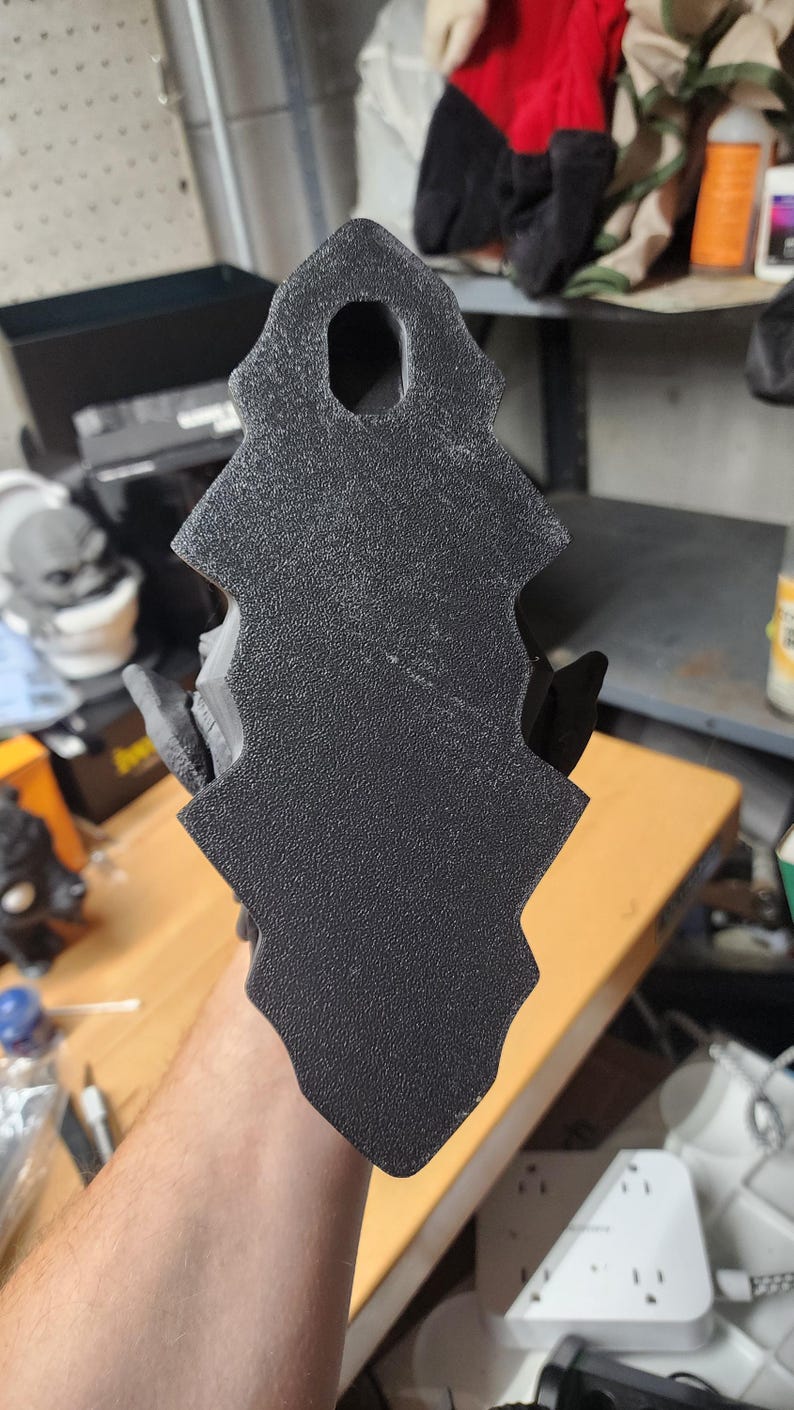 May include: A black, textured, vertically oriented object with a jagged, serrated edge profile and a single oval-shaped hole near the top. The surface has a matte, granular finish, and the overall shape is elongated with a pointed top and bottom.