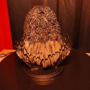 May include: A dark bronze-coloured bust sculpture depicting a figure with textured, shaggy hair and a layered, jagged collar. The piece is mounted on a circular base, showcasing intricate, rough detailing across the surface of the hair and clothing.
