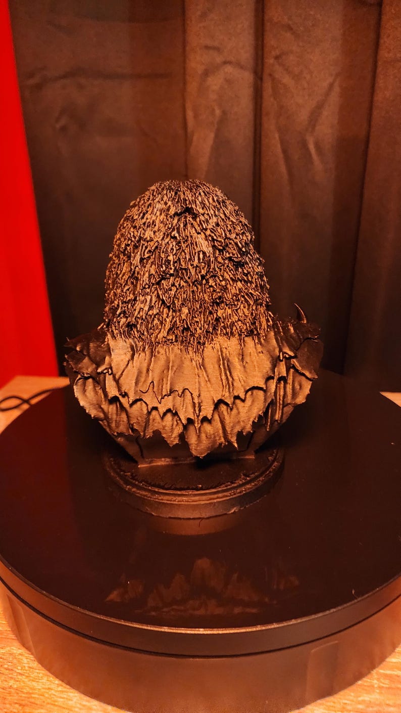 May include: A dark bronze-coloured bust sculpture depicting a figure with textured, shaggy hair and a layered, jagged collar. The piece is mounted on a circular base, showcasing intricate, rough detailing across the surface of the hair and clothing.