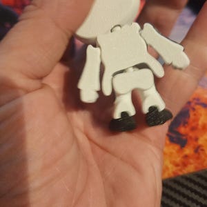 May include: A small, articulated white figurine with a rounded head, a torso, and limbs, featuring black feet. The figure has a textured surface and is constructed with visible joints at the shoulders, hips, and neck, allowing for movement.