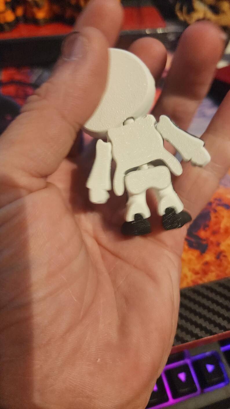 May include: A small, articulated white figurine with a rounded head, a torso, and limbs, featuring black feet. The figure has a textured surface and is constructed with visible joints at the shoulders, hips, and neck, allowing for movement.