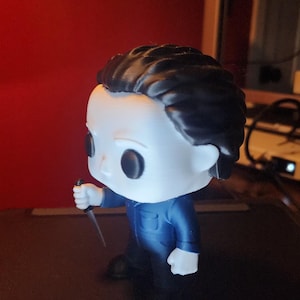 May include: A vinyl collectible figure featuring a character with a white mask, black hair, and a blue jumpsuit. The figure is holding a small silver-colored knife in its right hand. The character has large black eyes and a neutral facial expression.