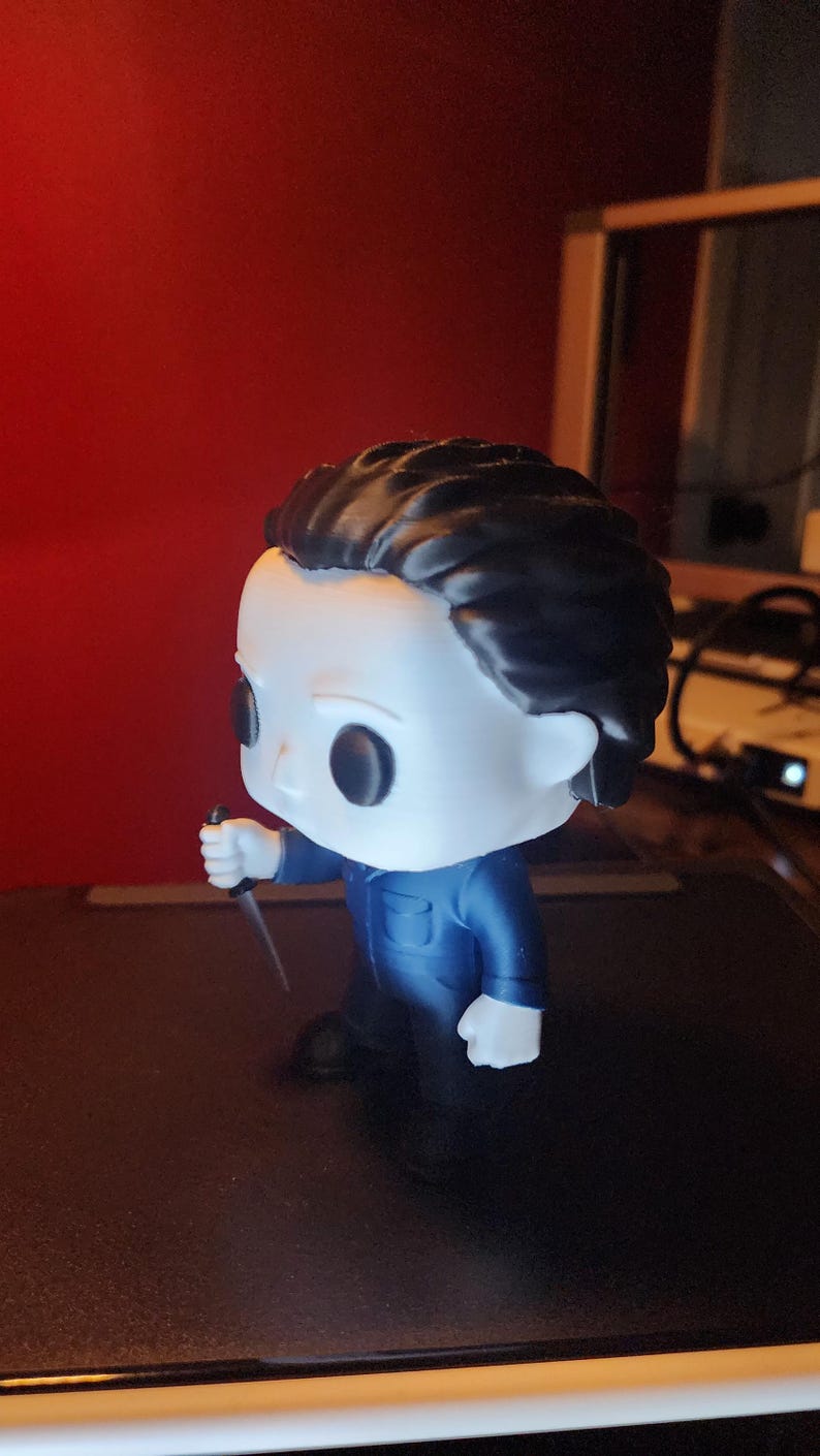 May include: A vinyl collectible figure featuring a character with a white mask, black hair, and a blue jumpsuit. The figure is holding a small silver-colored knife in its right hand. The character has large black eyes and a neutral facial expression.
