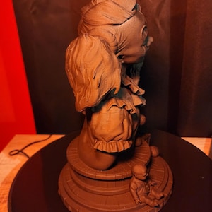 May include: A monochromatic brown sculpture of a clown figure with a ruffled collar and hair in pigtails, positioned on a circular base. A small, seated doll figure is placed on the edge of the base, which features a wood-plank texture.