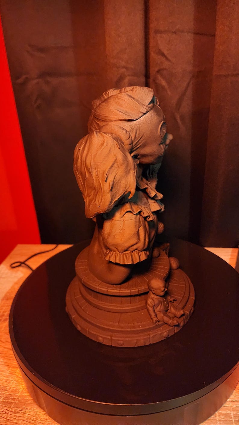 May include: A monochromatic brown sculpture of a clown figure with a ruffled collar and hair in pigtails, positioned on a circular base. A small, seated doll figure is placed on the edge of the base, which features a wood-plank texture.