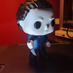 May include: A stylized vinyl figure of a character with a white mask, black hair, and a dark blue jumpsuit. The figure has large black circular eyes and is holding a small silver-colored knife in its right hand. The design features a large head.