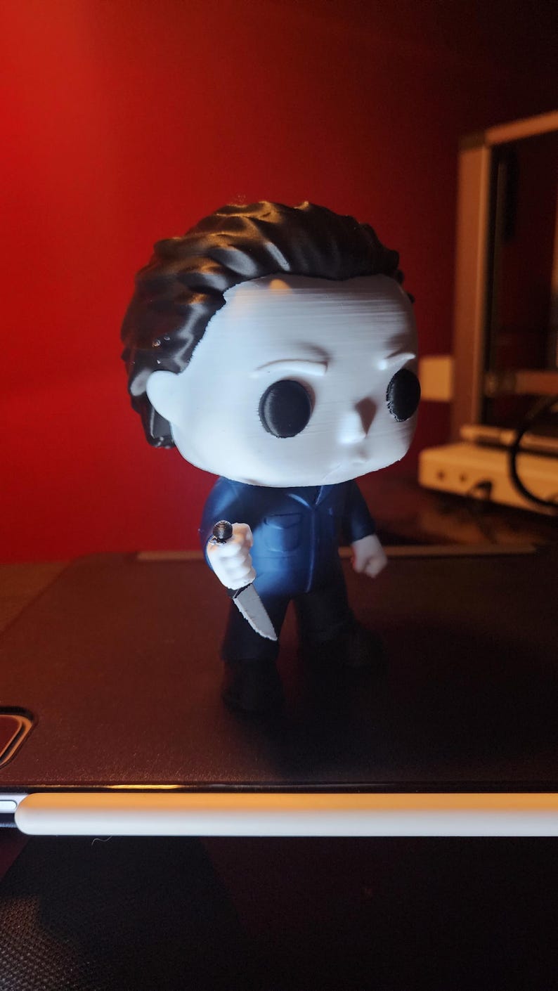 May include: A stylized vinyl figure of a character with a white mask, black hair, and a dark blue jumpsuit. The figure has large black circular eyes and is holding a small silver-colored knife in its right hand. The design features a large head.