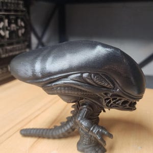 May include: A black vinyl collectible figure of a Xenomorph creature from the Alien franchise. The figure features an elongated, smooth head, a segmented body with visible ribbing, clawed hands, and a long, pointed tail extending behind it.