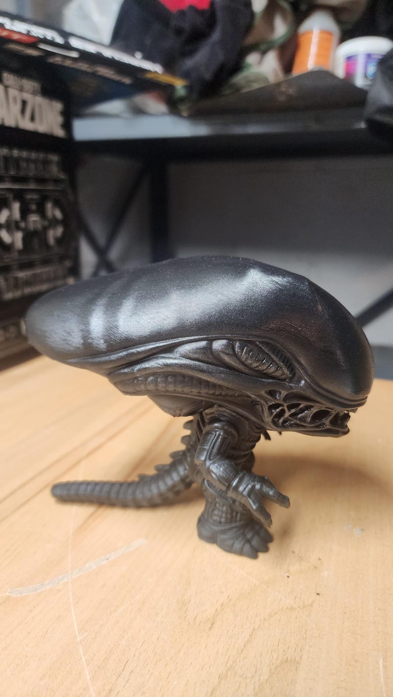 May include: A black vinyl collectible figure of a Xenomorph creature from the Alien franchise. The figure features an elongated, smooth head, a segmented body with visible ribbing, clawed hands, and a long, pointed tail extending behind it.