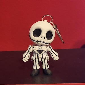 May include: A white and black character keychain featuring a round head with large hollow eyes and a stitched mouth. The figure wears a pinstriped suit with a bow tie and black shoes. A silver metal ring and clasp are attached to the top of the head.