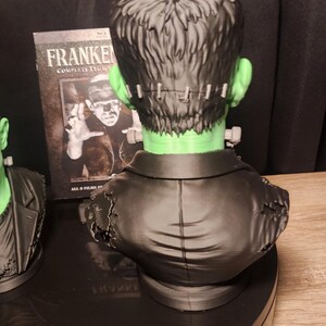 May include: A green-skinned Frankenstein monster bust featuring black hair, a stitched forehead, and a black jacket with distressed shoulder details. Behind the bust stands a Blu-ray case titled Frankenstein Complete Legacy Collection.