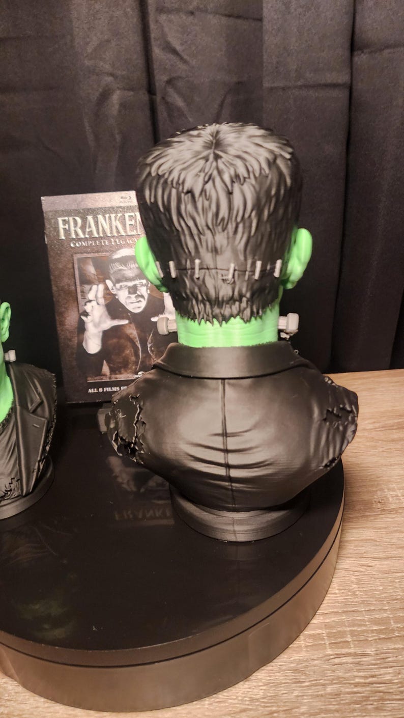 May include: A green-skinned Frankenstein monster bust featuring black hair, a stitched forehead, and a black jacket with distressed shoulder details. Behind the bust stands a Blu-ray case titled Frankenstein Complete Legacy Collection.