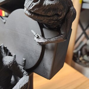 May include: A matte black 3D printed sculpture featuring a textured creature with a wrinkled face and webbed hands, perched on the edge of a toilet seat. The creature holds a small, rectangular object in one hand while resting on the lid.