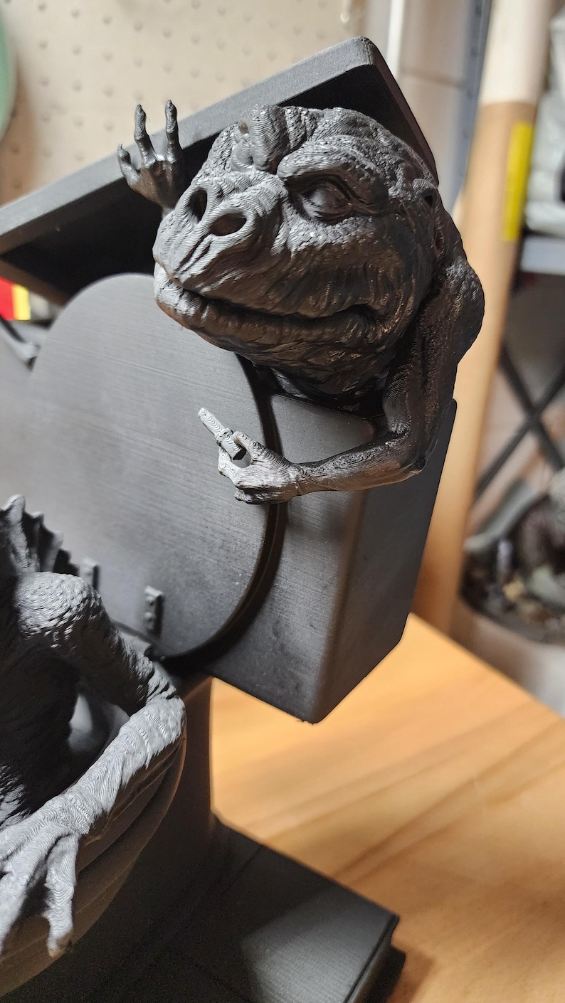May include: A matte black 3D printed sculpture featuring a textured creature with a wrinkled face and webbed hands, perched on the edge of a toilet seat. The creature holds a small, rectangular object in one hand while resting on the lid.