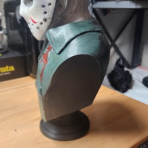 May include: A sculpted bust of a figure wearing a white hockey mask with red markings and a dark green collared shirt. The figure has a textured, pale gray head and a small red blood-like stain on the chest area, mounted on a black circular base.