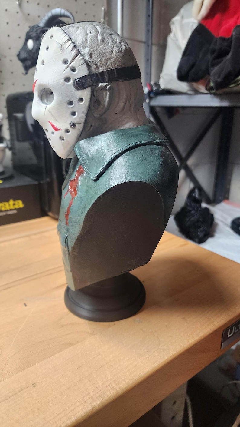 May include: A sculpted bust of a figure wearing a white hockey mask with red markings and a dark green collared shirt. The figure has a textured, pale gray head and a small red blood-like stain on the chest area, mounted on a black circular base.
