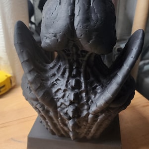 Puede incluir: Un busto esculpido en impresi&oacute;n 3D de color negro mate que presenta un cr&aacute;neo org&aacute;nico muy texturizado similar a un cerebro, una estructura vertebral con crestas prominentes y ap&eacute;ndices en forma de alas a los lados, montado sobre una base cuadrada.