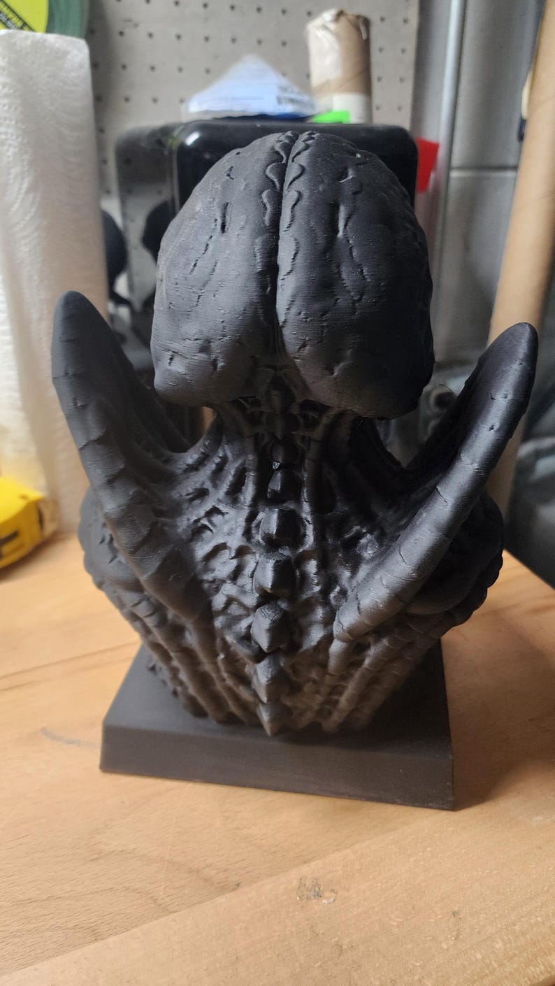 Puede incluir: Un busto esculpido en impresi&oacute;n 3D de color negro mate que presenta un cr&aacute;neo org&aacute;nico muy texturizado similar a un cerebro, una estructura vertebral con crestas prominentes y ap&eacute;ndices en forma de alas a los lados, montado sobre una base cuadrada.