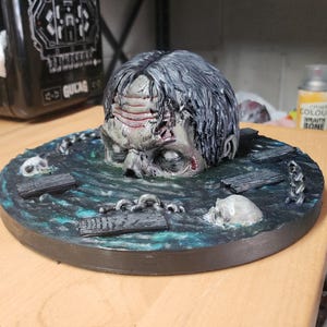 May include: A hand-painted figurine featuring a human head emerging from a dark, textured water surface. The base includes sculpted details of wooden planks, metal chains, and two skulls. The head has grey hair, visible forehead wrinkles, and pale, blank eyes, set against a circular, dark blue and green painted base.