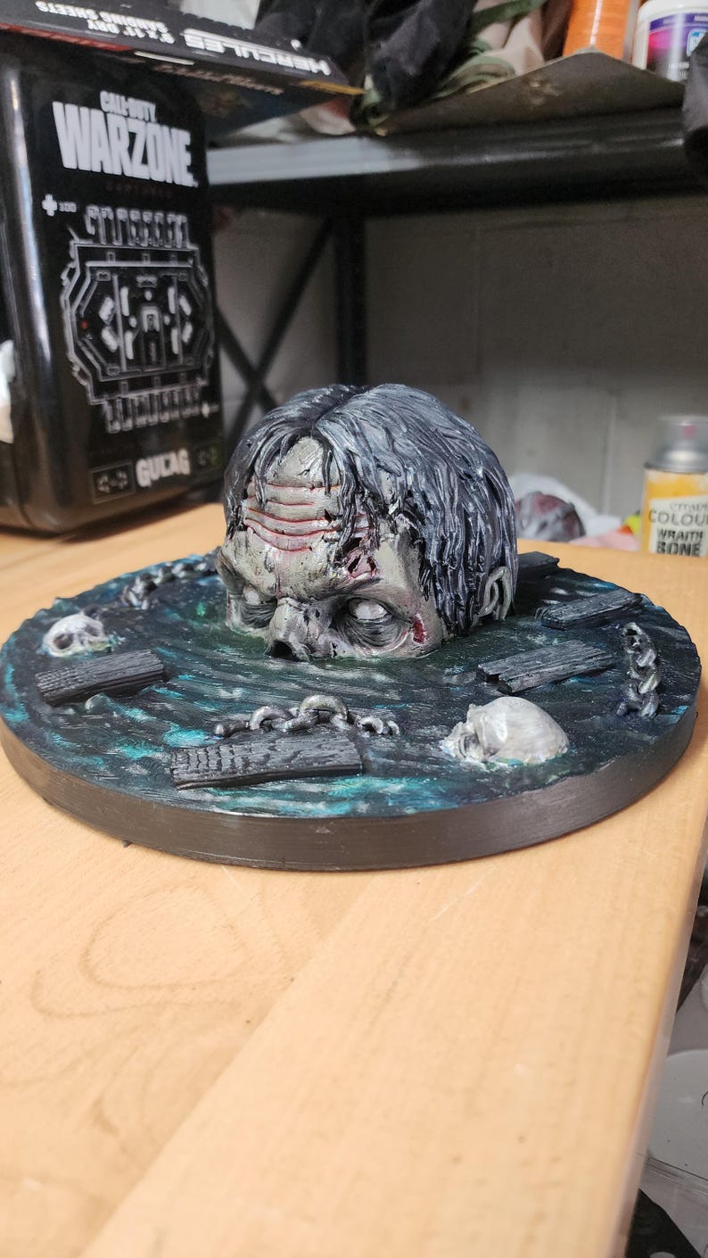 May include: A hand-painted figurine featuring a human head emerging from a dark, textured water surface. The base includes sculpted details of wooden planks, metal chains, and two skulls. The head has grey hair, visible forehead wrinkles, and pale, blank eyes, set against a circular, dark blue and green painted base.
