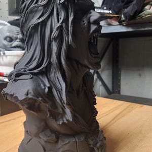 May include: A matte black bust sculpture of a figure with long, flowing hair and an open mouth revealing teeth. The figure has a rugged, textured appearance with a tattered garment around the shoulders and a circular base with cracked detailing.