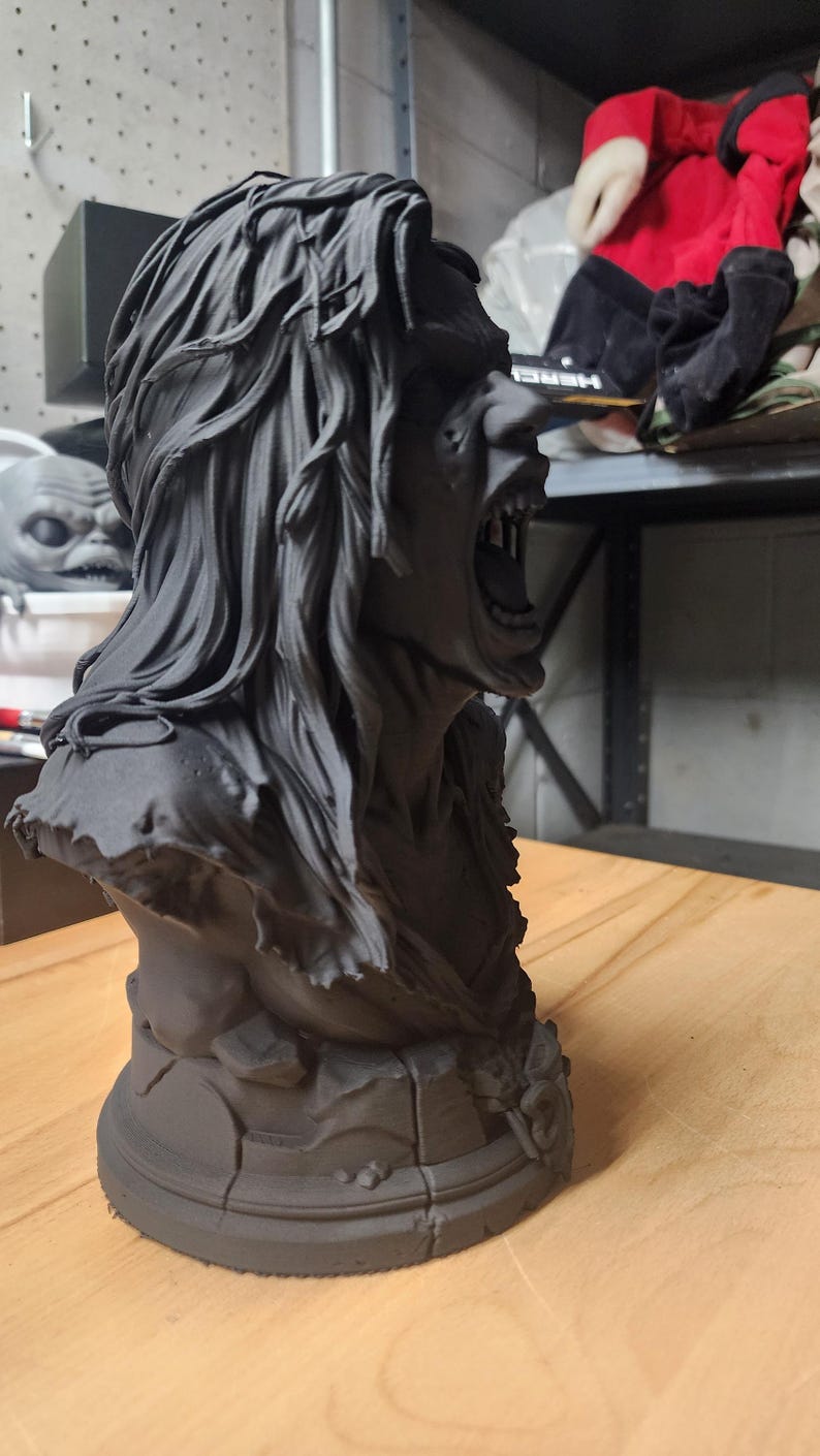 May include: A matte black bust sculpture of a figure with long, flowing hair and an open mouth revealing teeth. The figure has a rugged, textured appearance with a tattered garment around the shoulders and a circular base with cracked detailing.