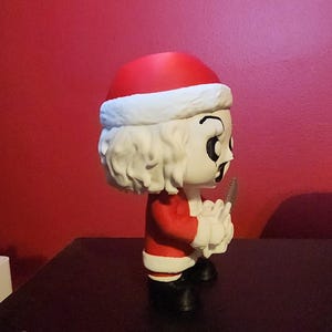 May include: A stylized vinyl figure of a character wearing a red and white Santa Claus suit with a matching hat. The figure has white hair, a pale face with dark eye makeup, and is holding a small gray serrated knife in its hands while wearing black boots.
