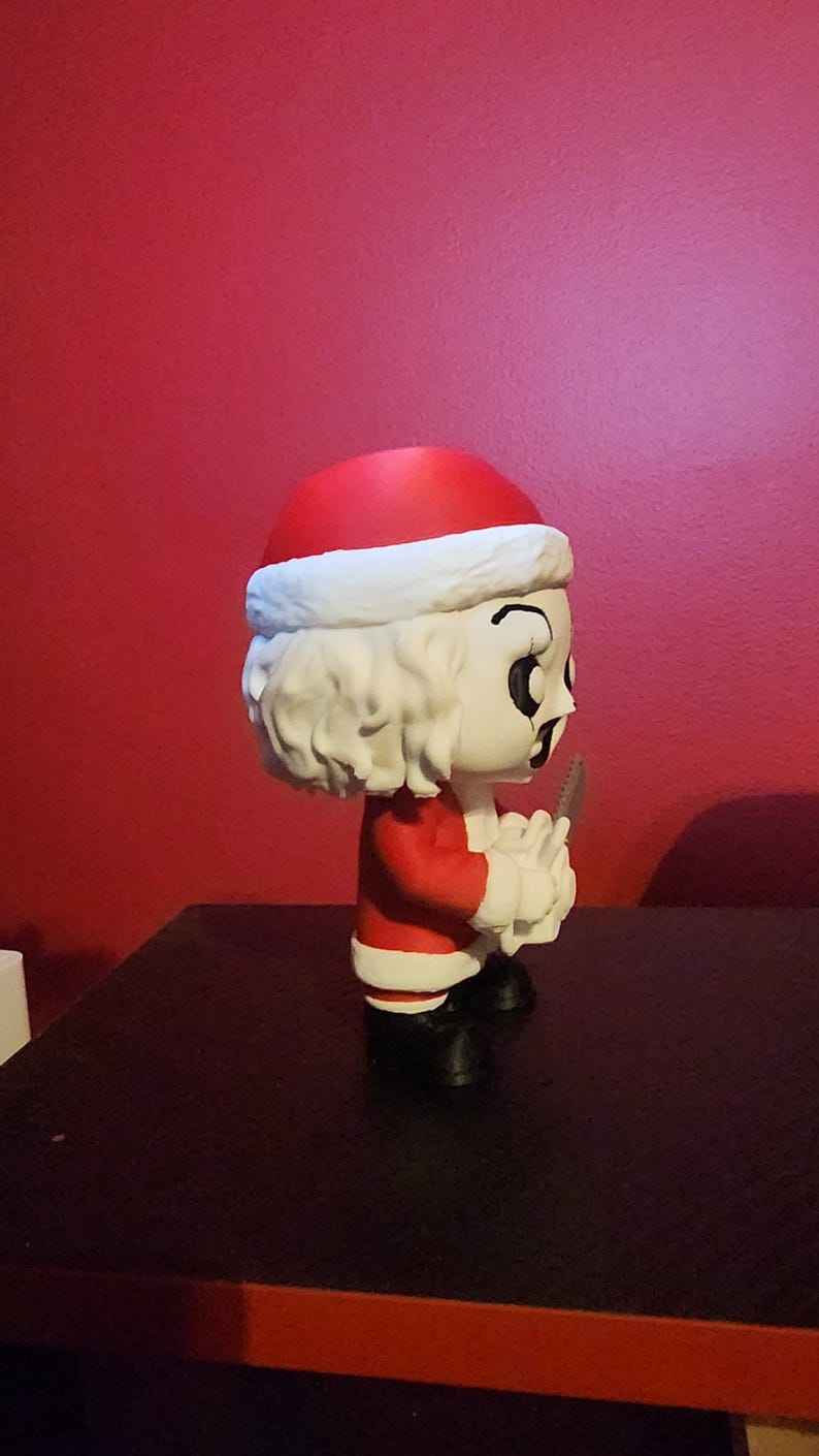 May include: A stylized vinyl figure of a character wearing a red and white Santa Claus suit with a matching hat. The figure has white hair, a pale face with dark eye makeup, and is holding a small gray serrated knife in its hands while wearing black boots.