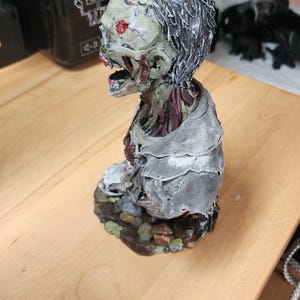 Op de afbeelding: Een handbeschilderd zombie-bustebeeldje met een bleekgroene huid, blootliggend rood spierweefsel en donker haar. Het figuur draagt een gehavend grijs kledingstuk en is gemonteerd op een basis van bruine en groene rotsen, met een open mond.