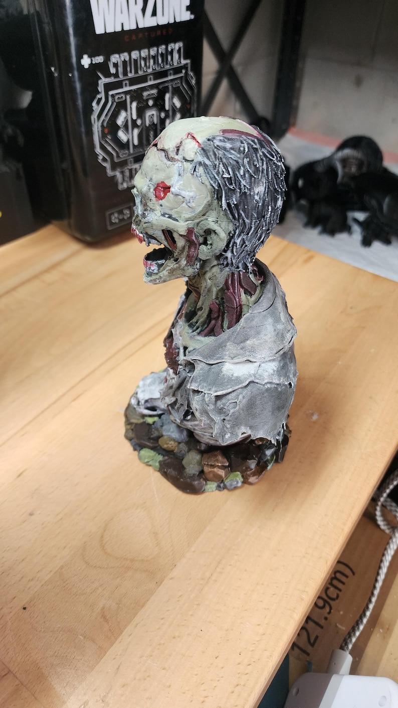 Op de afbeelding: Een handbeschilderd zombie-bustebeeldje met een bleekgroene huid, blootliggend rood spierweefsel en donker haar. Het figuur draagt een gehavend grijs kledingstuk en is gemonteerd op een basis van bruine en groene rotsen, met een open mond.