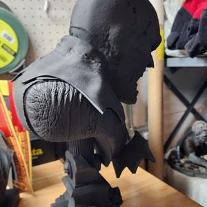 May include: A matte black 3D printed bust of a caped superhero character with pointed ears, textured skin, and a flowing cape. The figure is mounted on a detailed architectural base featuring small columns and structures, presented in a side profile view.