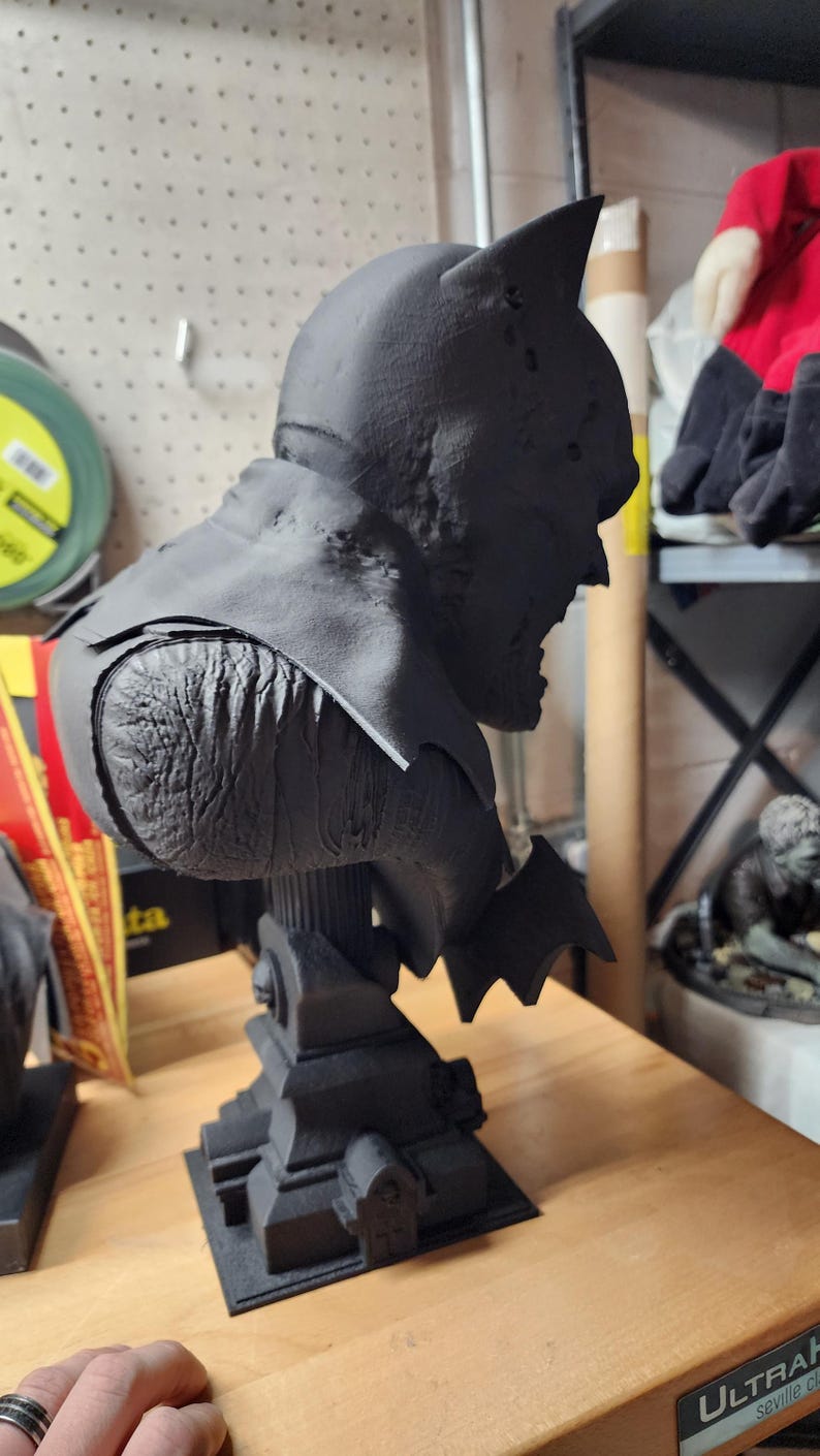 May include: A matte black 3D printed bust of a caped superhero character with pointed ears, textured skin, and a flowing cape. The figure is mounted on a detailed architectural base featuring small columns and structures, presented in a side profile view.