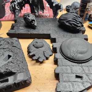 May include: A collection of unpainted black miniature gaming terrain pieces, including a large creature figure, a skull, a textured rock, and stone-patterned floor tiles. These plastic components feature detailed surfaces and are ready for assembly.