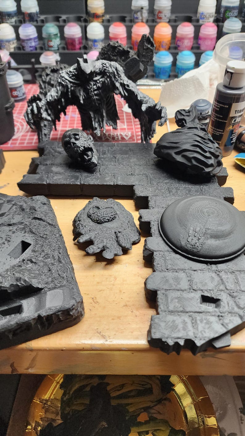 May include: A collection of unpainted black miniature gaming terrain pieces, including a large creature figure, a skull, a textured rock, and stone-patterned floor tiles. These plastic components feature detailed surfaces and are ready for assembly.