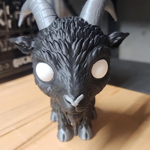 May include: A black goat figurine with large, smooth grey horns, a textured coat, and solid white circular eyes. The goat has a small grey nose and grey hooves, featuring a stylized, oversized head design with a pointed beard under the chin.