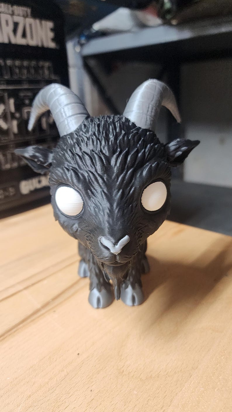 May include: A black goat figurine with large, smooth grey horns, a textured coat, and solid white circular eyes. The goat has a small grey nose and grey hooves, featuring a stylized, oversized head design with a pointed beard under the chin.