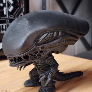 May include: A matte black figurine of a Xenomorph creature from the Alien franchise. The figure features an elongated, smooth head, a detailed skeletal torso, sharp claws, and a long tail. It stands in a crouched position with visible teeth.