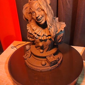 May include: A monochromatic dark brown bust of a clown with a ruffled collar, featuring a small doll figure and a square box with a star design on the circular base. The clown has detailed facial features, messy hair, and a wide, expressive smile.