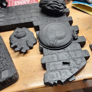 May include: A black 3D printed miniature terrain piece featuring a circular stone well cover set into a textured brick wall section, accompanied by a separate small black sculpted creature figure and a larger black jagged rock formation.