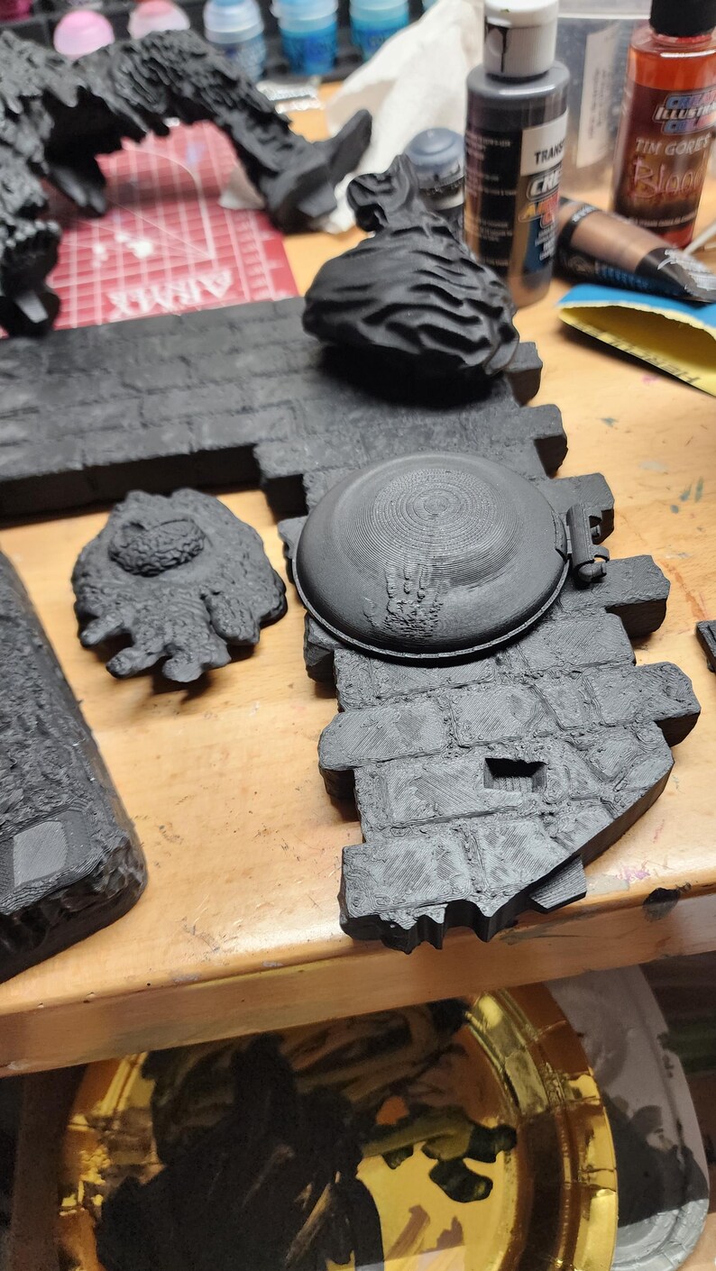 May include: A black 3D printed miniature terrain piece featuring a circular stone well cover set into a textured brick wall section, accompanied by a separate small black sculpted creature figure and a larger black jagged rock formation.