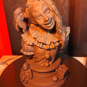 May include: A monochromatic brown bust of a clown with a wide, toothy grin, wearing a ruffled collar and a shirt with two spherical buttons. The base features a small doll figure, a crate filled with balls, and a single ball resting on the surface.