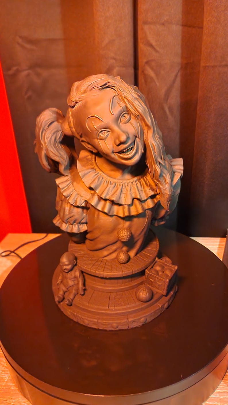 May include: A monochromatic brown bust of a clown with a wide, toothy grin, wearing a ruffled collar and a shirt with two spherical buttons. The base features a small doll figure, a crate filled with balls, and a single ball resting on the surface.